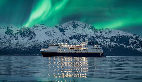 Expedition cruise ship sailing in front of snow-covered mountains beneath the northern lights.
