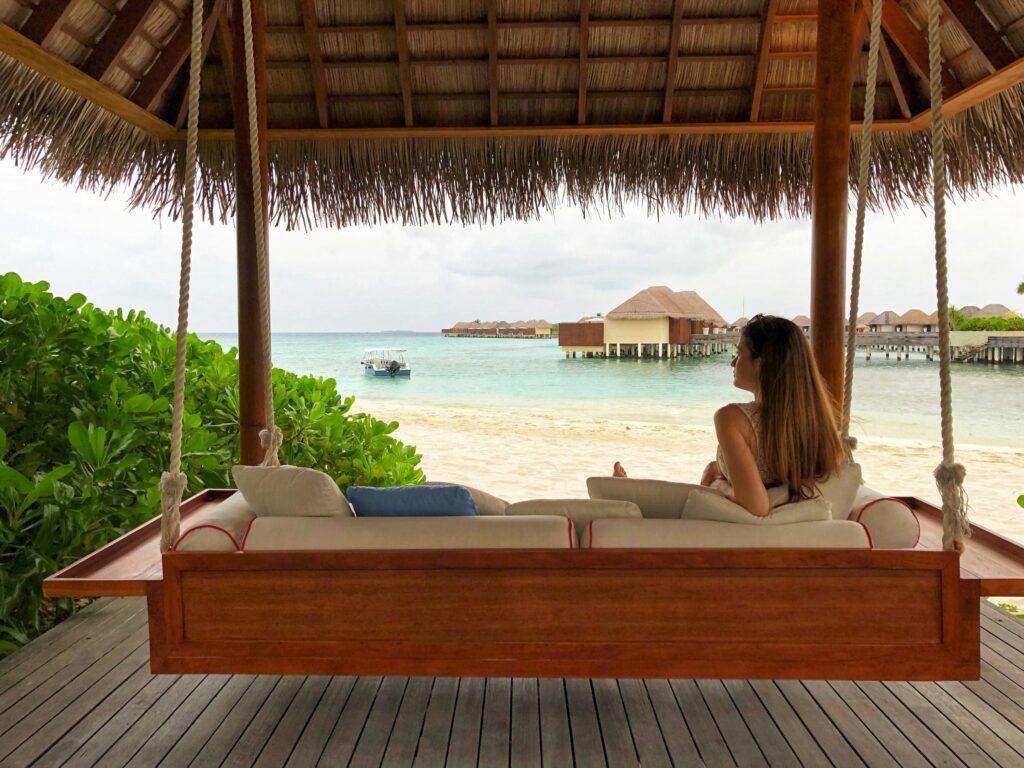 Woman sitting on a wooden swing bed under a thatched roof, overlooking a beach and overwater villas.