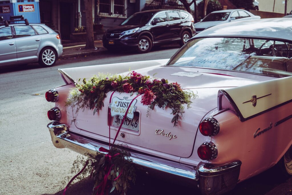 Vintage pink Dodge with a “Just Married” sign and floral arrangement on the back, parked along a residential street.